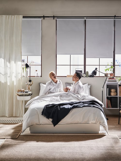 Bedroom with two people sitting in a bed frame with storage, dressed in a  duvet cover, with a throw, and a pair of curtains.