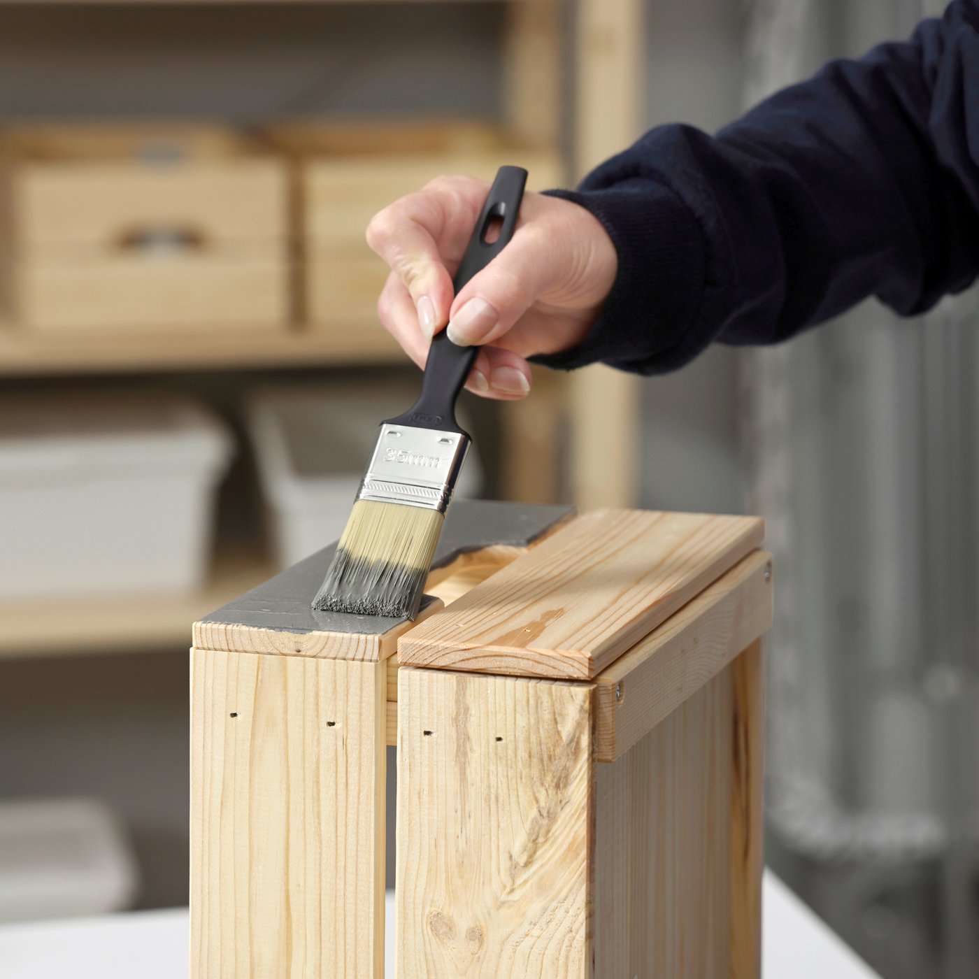 Person streicht die obere Kante einer Holzkiste mit einem Pinsel & grauem TRIXIG-Lack; im Hintergrund Regale mit verschiedenen Gegenständen.