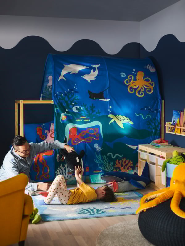 A man dangles a BLÅVINGAD orca soft toy above a child lying on a BLÅVINGAD rug beside a bed covered in a KURA bed tent.