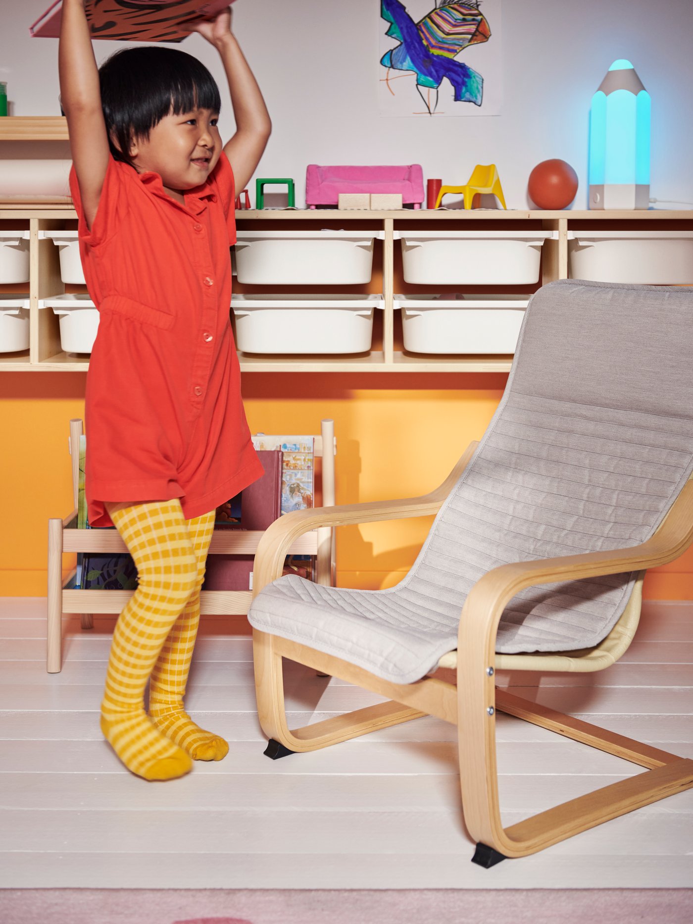 A child holding a book over their head in front of a FLISAT book display, and facing towards a light beige POÄNG armchair.