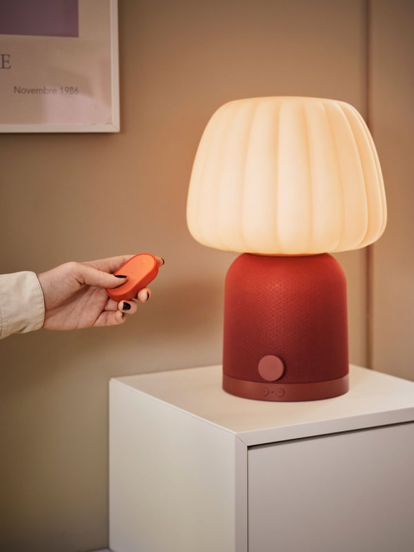 En hand håller en liten BILRESA fjärrkontroll framför en bordslampa med varm belysning och röd fot, placerad på ett skåp bredvid en växt.