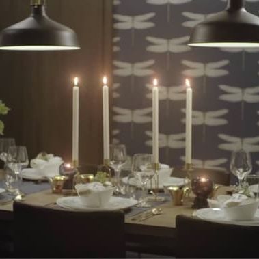 Festlich gedeckter Esstisch mit Kerzen, weissem Geschirr, Gläsern und goldenen Akzenten unter schwarzen Pendelleuchten.