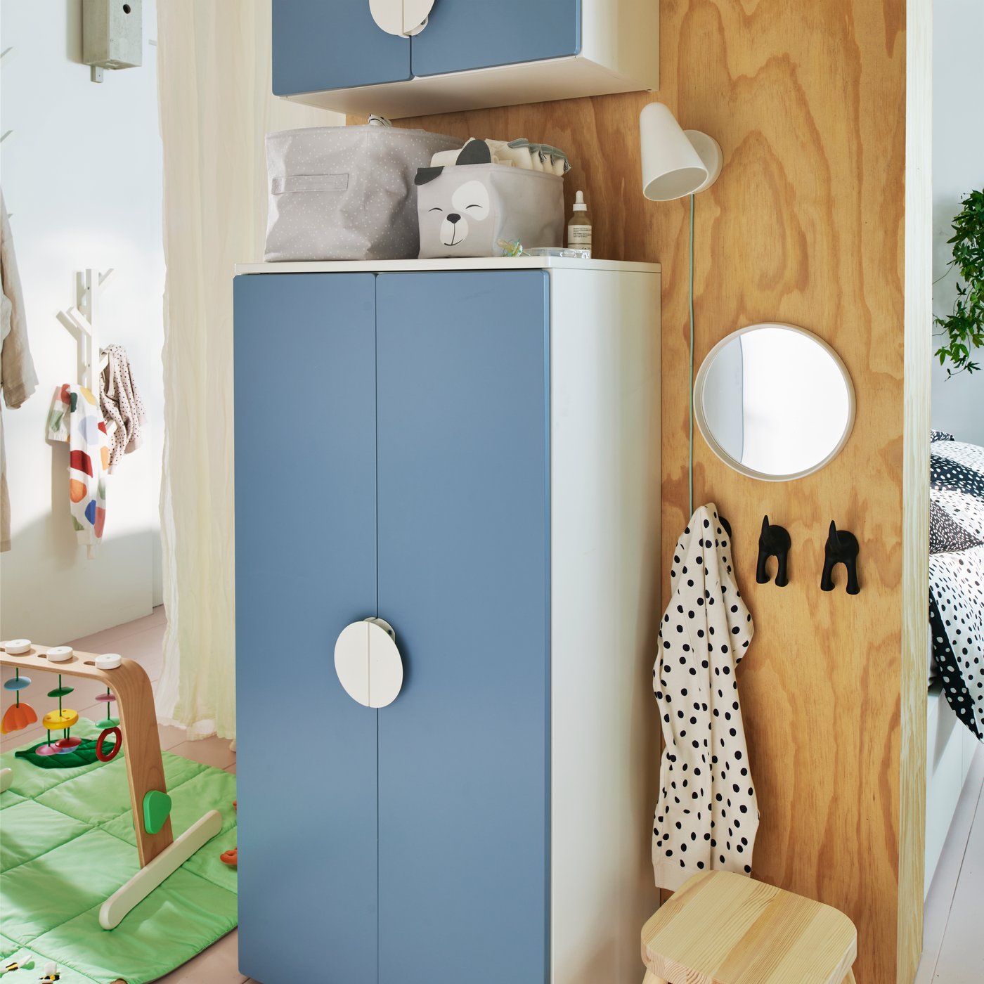 A blue SMÅSTAD wardrobe against a wooden wall with two storage boxes holding baby things on top and a wall light beside it.