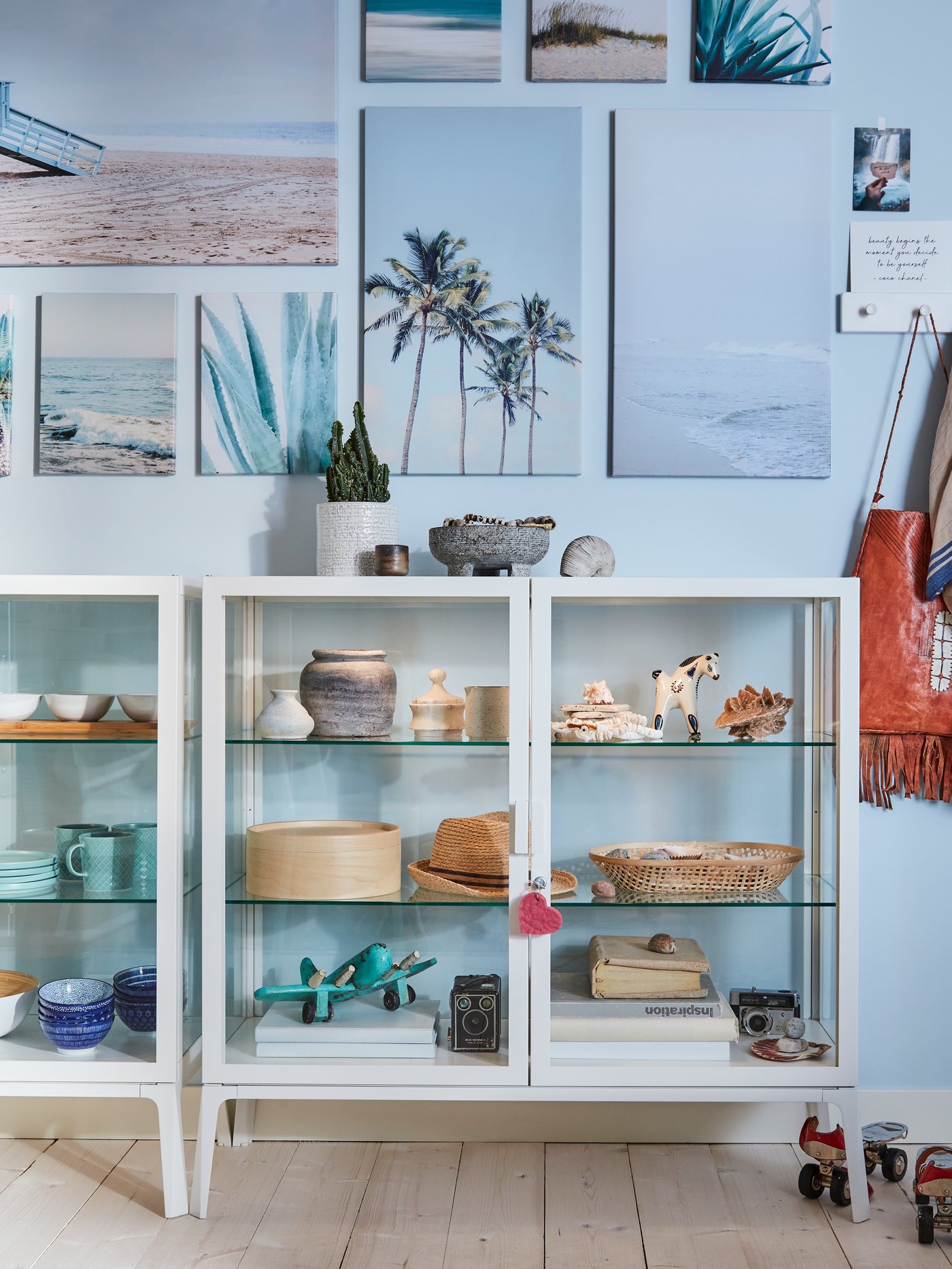 Dua kabinet pintu kaca berwarna putih dengan unit hiasan di dalam, dinding berwarna biru dan cetakan pantai digantung pada dinding.