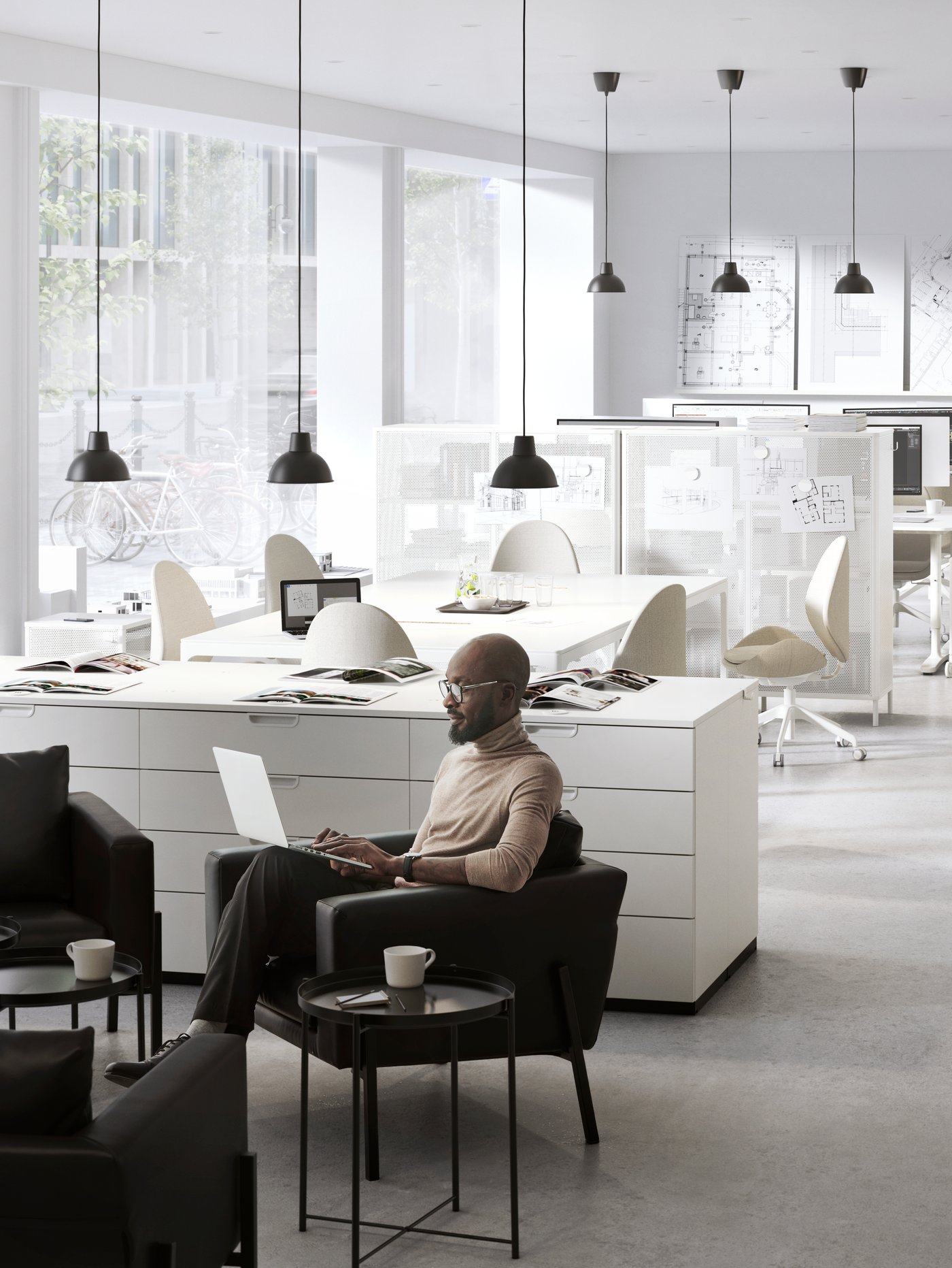 Espacio de trabajo con cajoneras blancas, lámparas de techo negras, sillas de oficina, una mesa de reuniones y un hombre sentado en un sillón.