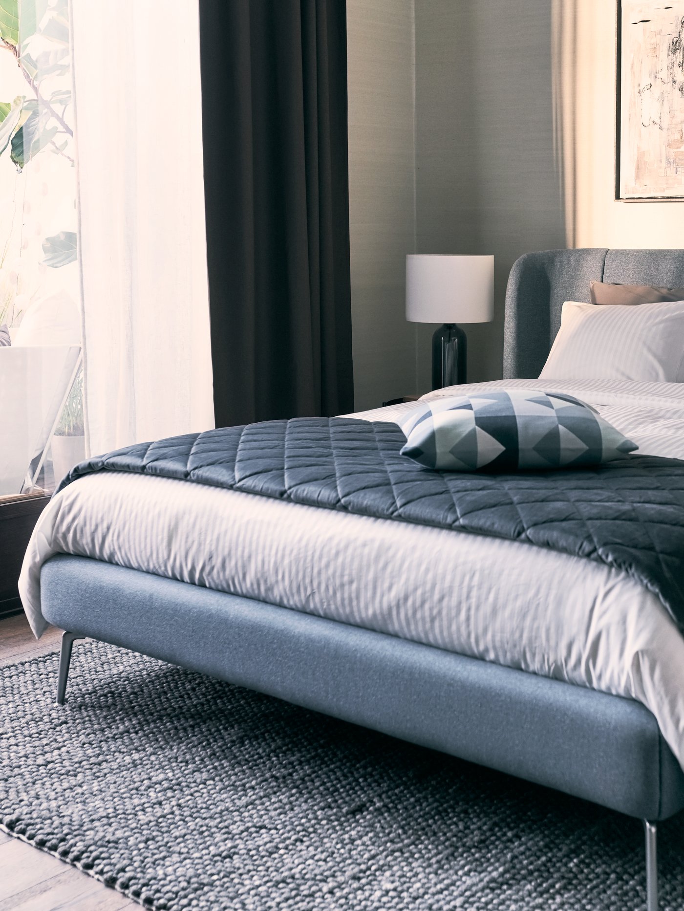 Modrá čalouněná postel TUFJORD s bílým povlečením NATTJASMIN, s tmavě šedým přehozem MJUKPLISTER a polštářem nahoře.