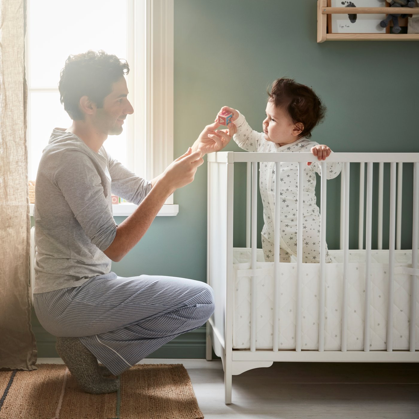 Baby standing in a white crib holding his father's finger that is squatting outside the crib to be eye level with his son.