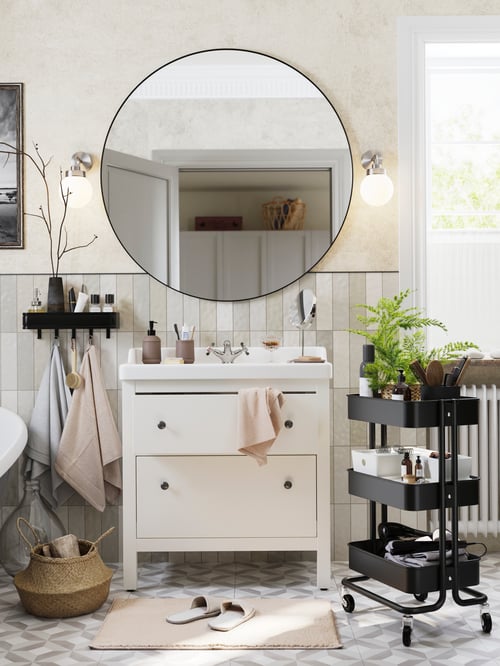 O baie cu o lavoar HEMNES cu sertare și o oglindă deasupra ei, împreună cu un cărucior negru și un coș pe fiecare parte.
