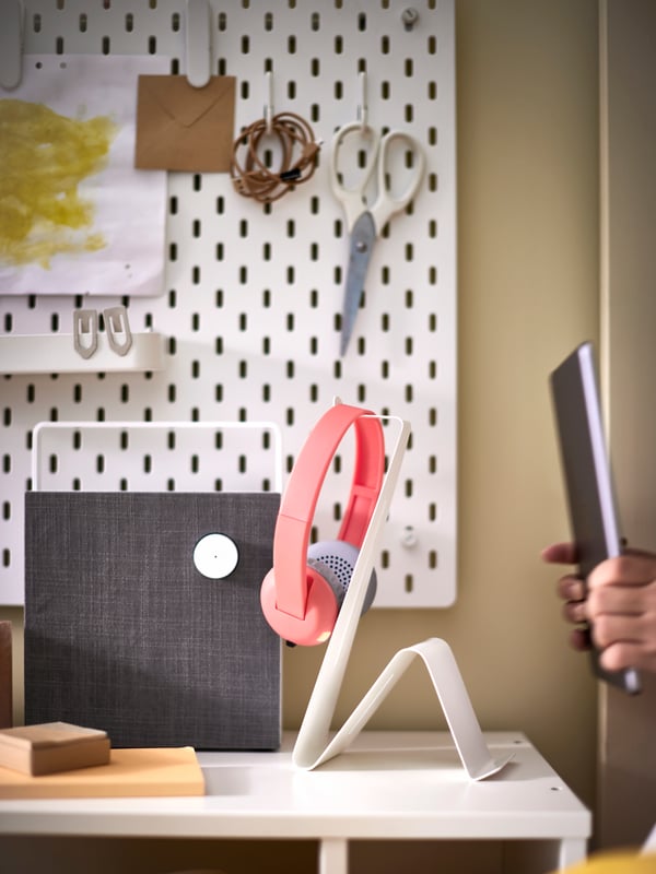 En el primer plano se ve un soporte para aparatos electrónicos MÖJLIGHET sobre una mesa blanca. En el muro del fondo, un tablero perforado SKÅDIS blanco con papeles y tijeras colgadas. 