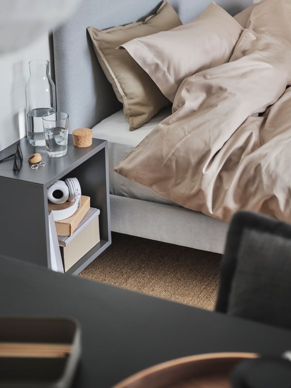 An upholstered bed with bedding in different beige colours, a wall-mounted bedside table with a jug of water and a glass.