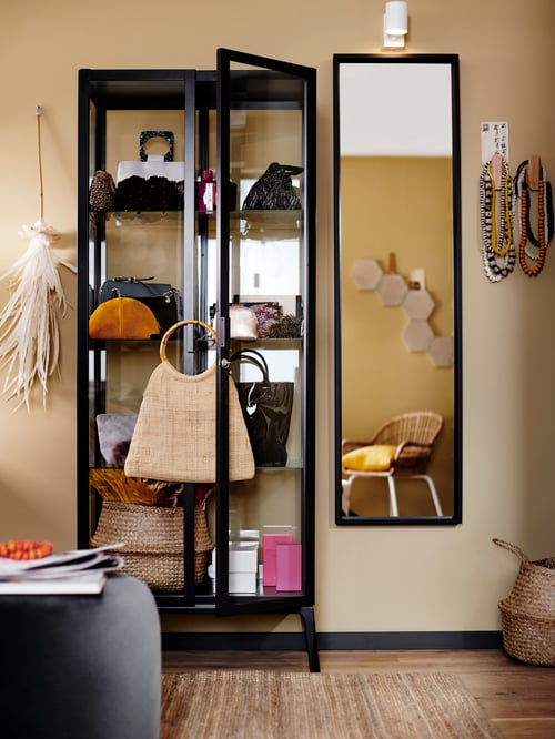 Bedroom with cream brown wall, glass-door cabinet with bags, boxes, pouffe, accessories, and mirror.