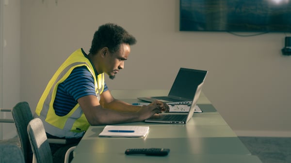 Abdi, wearing a shirt and safety vest, is focused on his laptop at a conference table, surrounded by a notebook and remote control.