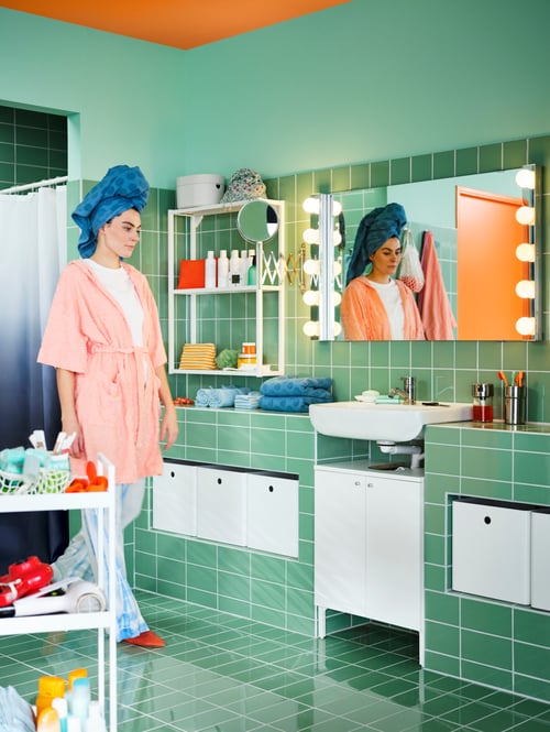 Ein grün gefliestes Badezimmer mit einem weißen BJÖRKÅN Waschbecken und einem NYSJÖN Waschbeckenunterschrank, einem LETTAN Spiegel und einer Frau im Schlafanzug.