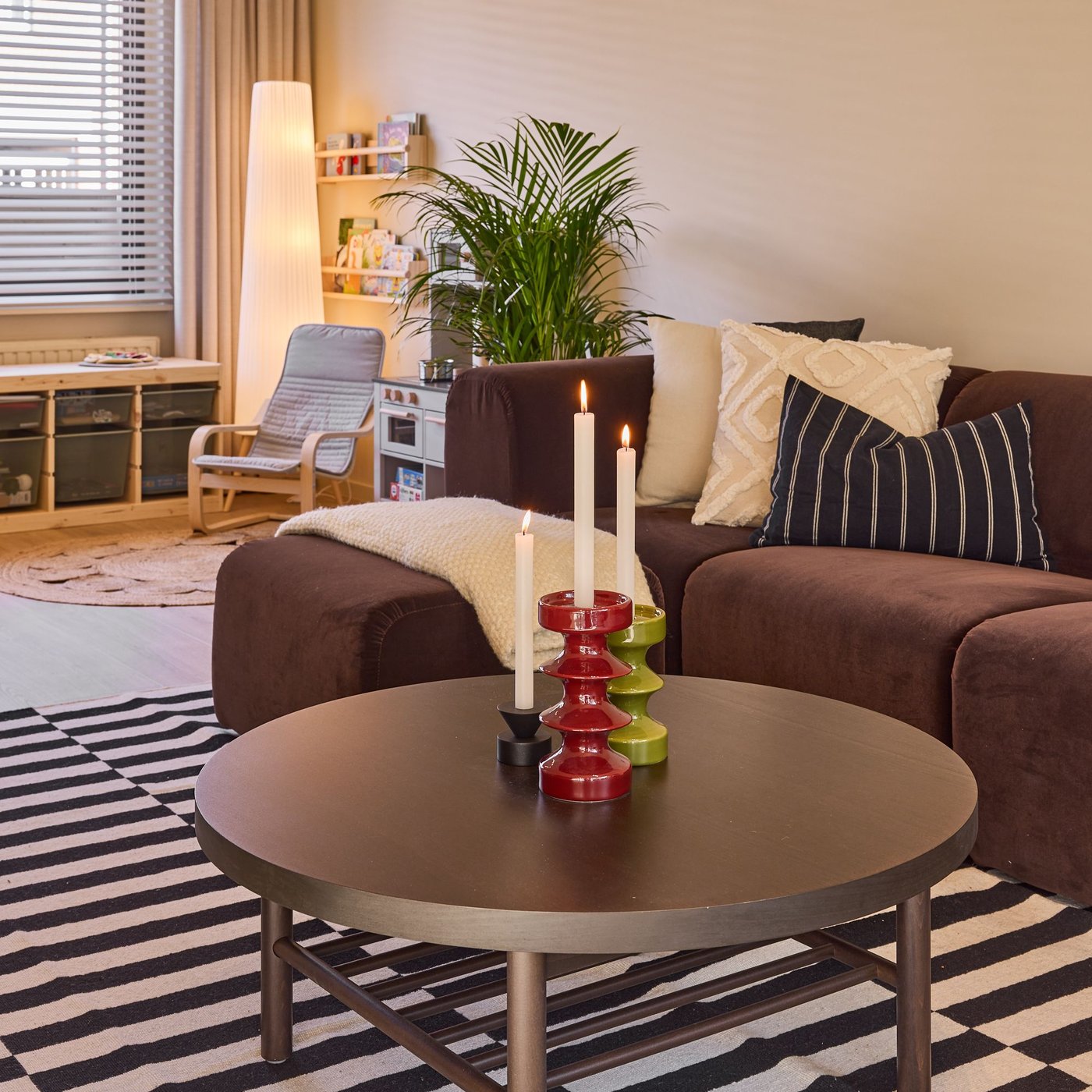 Ronde salontafel met kandelaars voor een bruine zetel met kussens, in een gezellige woonkamer met tapijt en planten.