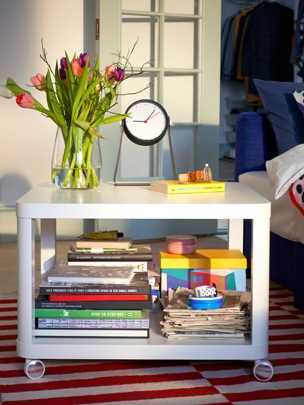TINGBY white side table on castors, with lots of books on the lower level and with a vase of flowers and a clock on top.