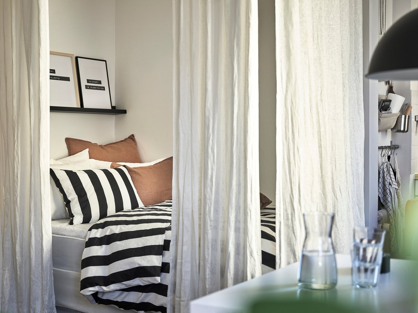 En säng med svart och vit randiga påslakan omringad av vita gardiner i en liten etta. I förgrunden syns en vattenkaraff och ett glas.