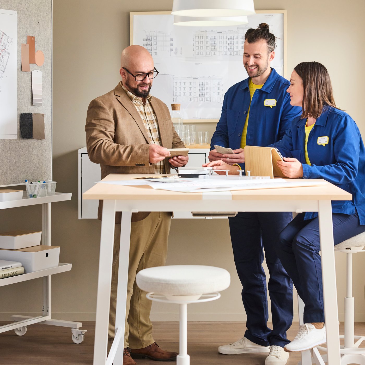 Tre kollegor runt ett högt bord granskar papper tillsammans, med en whiteboardtavla, hyllor och kontorsverktyg i bakgrunden.