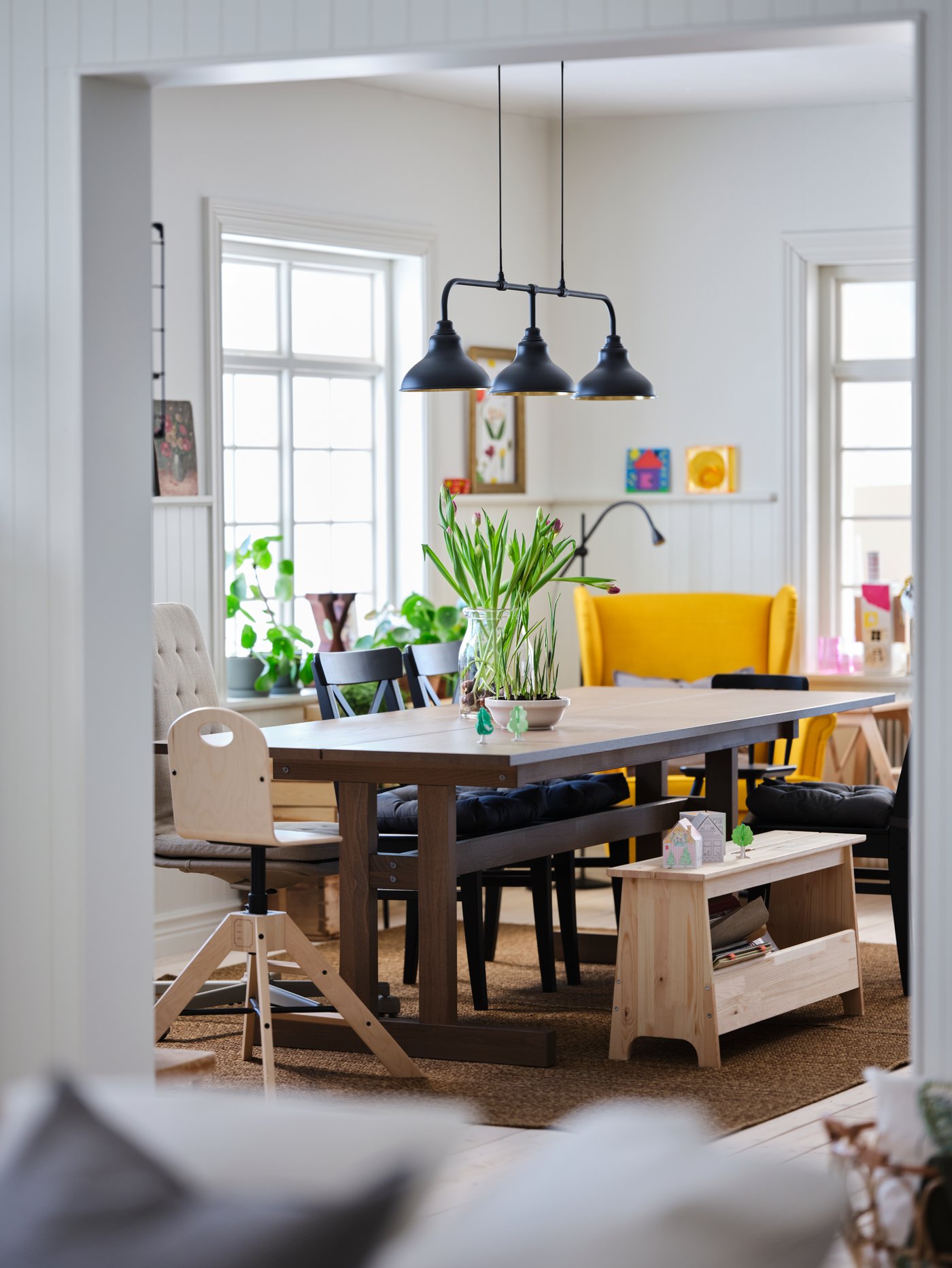 Mesa de comedor KLIMPFJÄLL con sillas de madera GRÅVAL y un sillón amarillo STRANDMON. Sobre ella hay 3 lámparas negras colgantes AGUNNARYD.