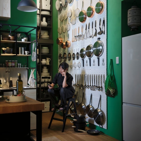 A person is sitting on a stepladder in front of a wall adorned with SKÅDIS pegboards, with kitchen items attached to them.