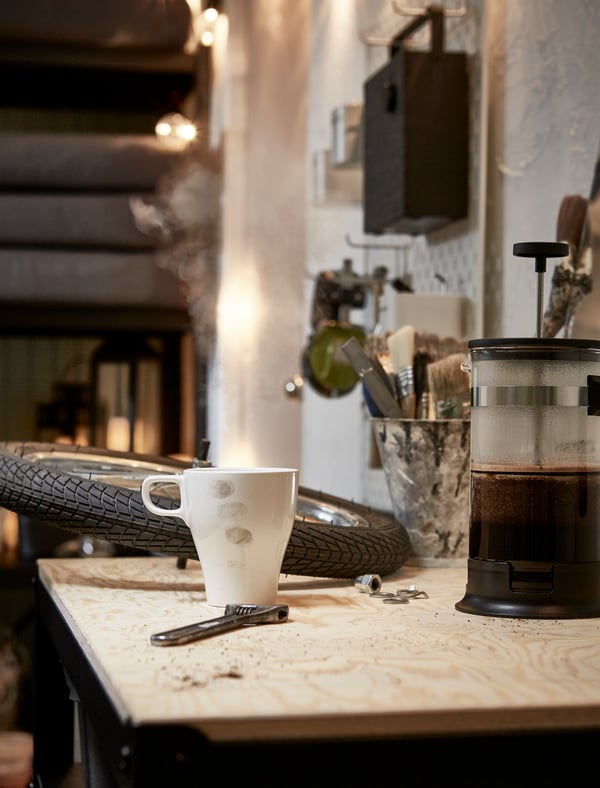 Arbejdsbord i et garagelignende rum med et cykelhjul og en skruenøgle, der deler bordpladsen med en stempelkande og et kaffekrus med fingeraftryk på.