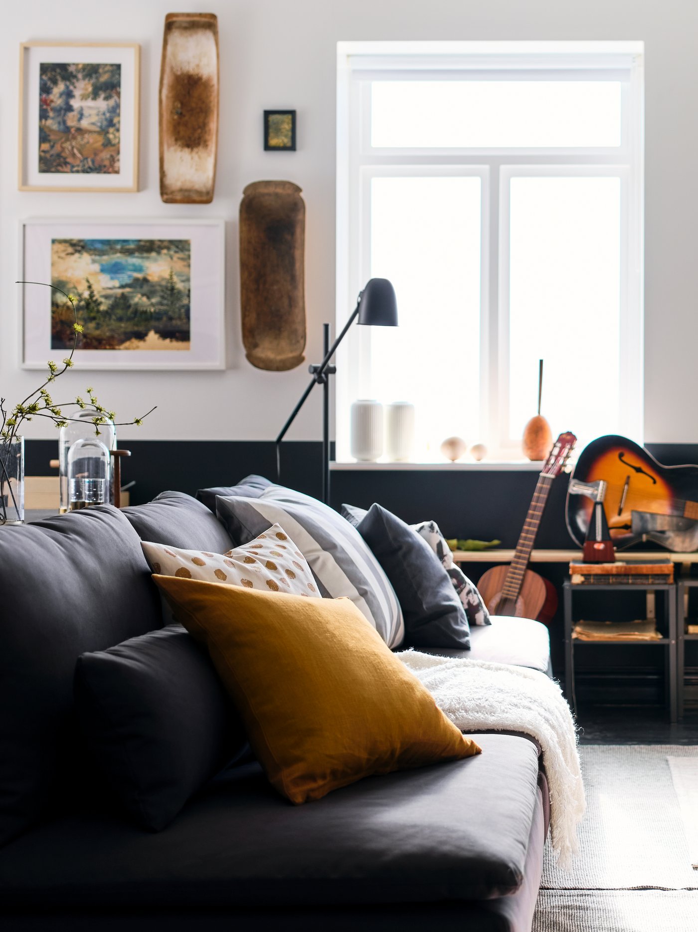 A section of a living room with a dark-grey SÖDERHAMN sofa, wall art, musical instruments and a black SKURUP reading lamp.