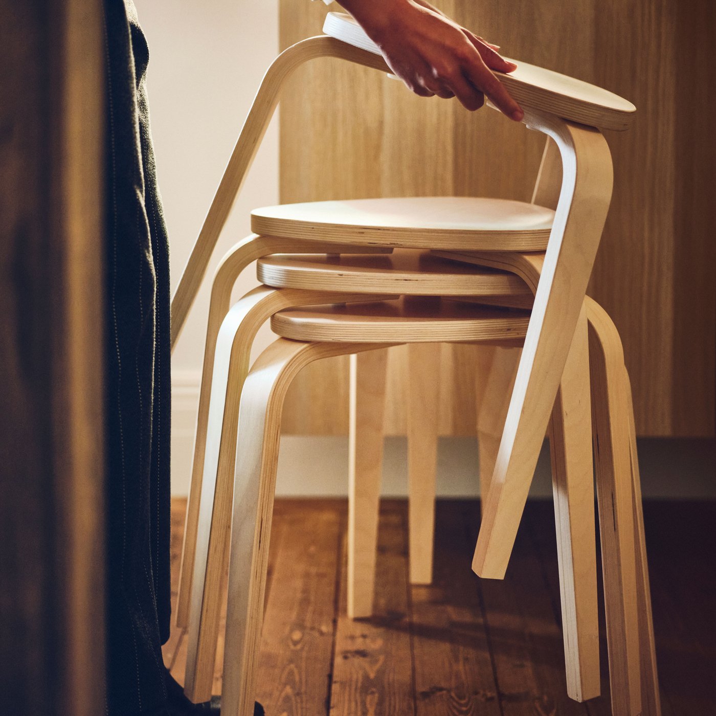 A person stacks another birch KYRRE stool on top of a stack of three other KYRRE stools in a room with a wooden floor.