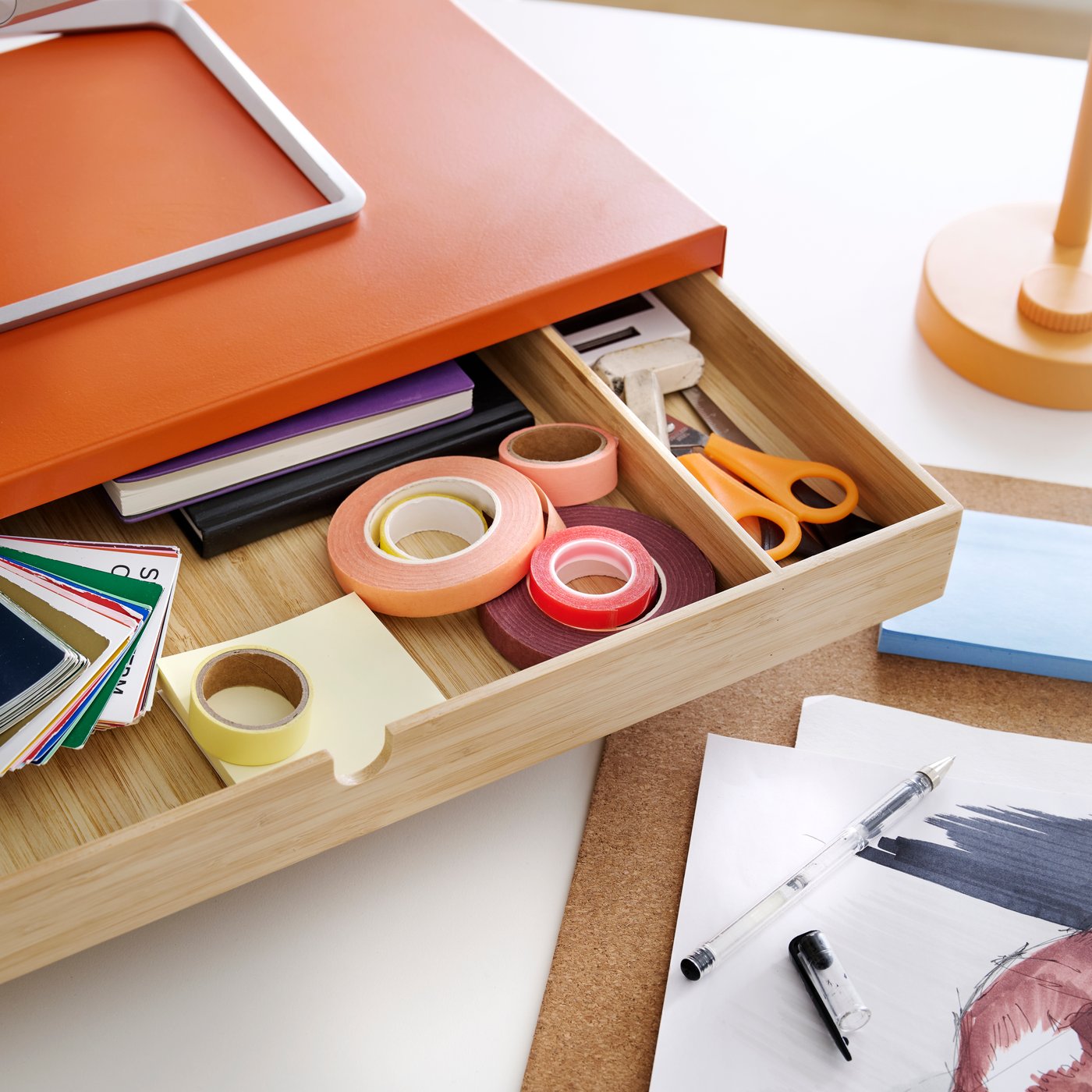 A close-up of a bright orange ELLOVEN monitor stand with drawer, holding stationery items on a desk.