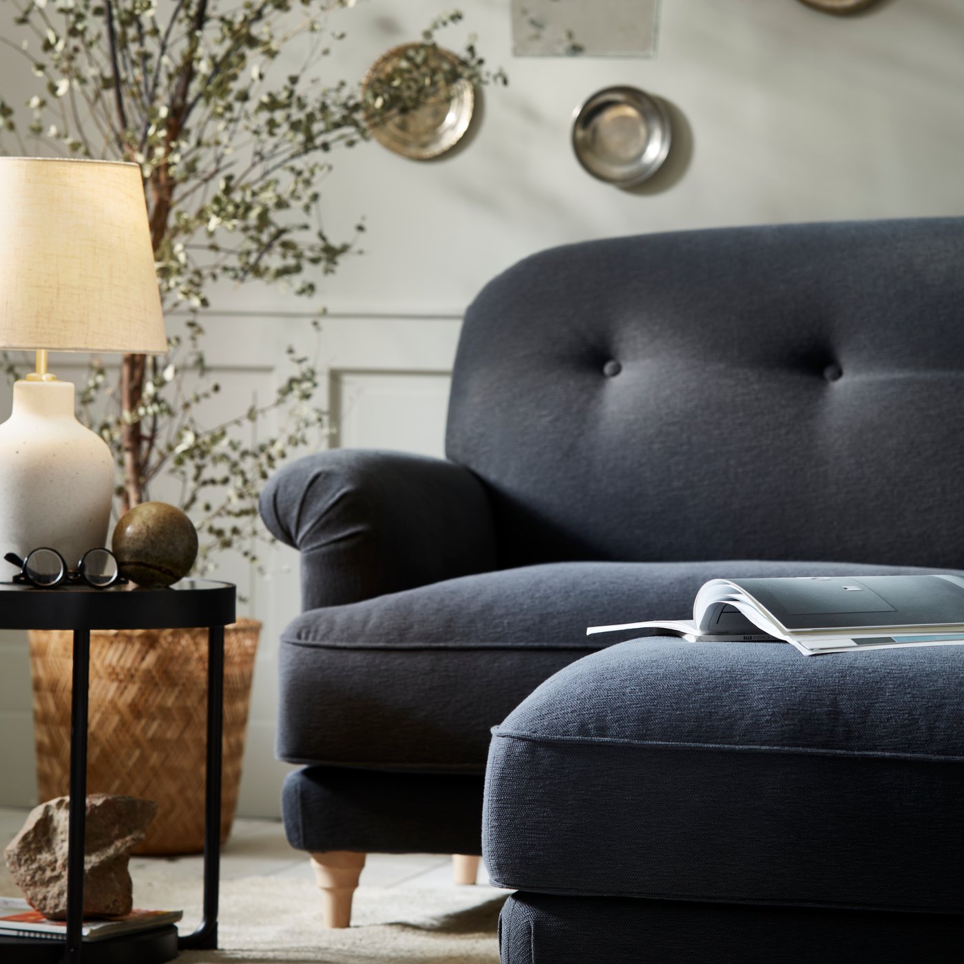 I bilden syns en halv mörkgrå soffa i klassisk stil och bredvid står ett litet svart sidobord med en bordslampa på.