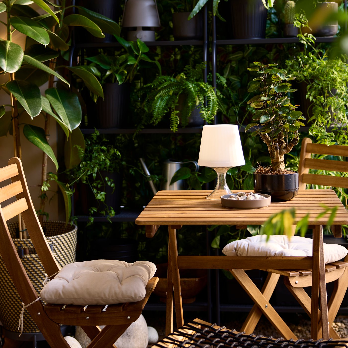 A light-brown stained acacia ASKHOLMEN foldable table and two folding chairs are on a balcony with shelves and potted plants.