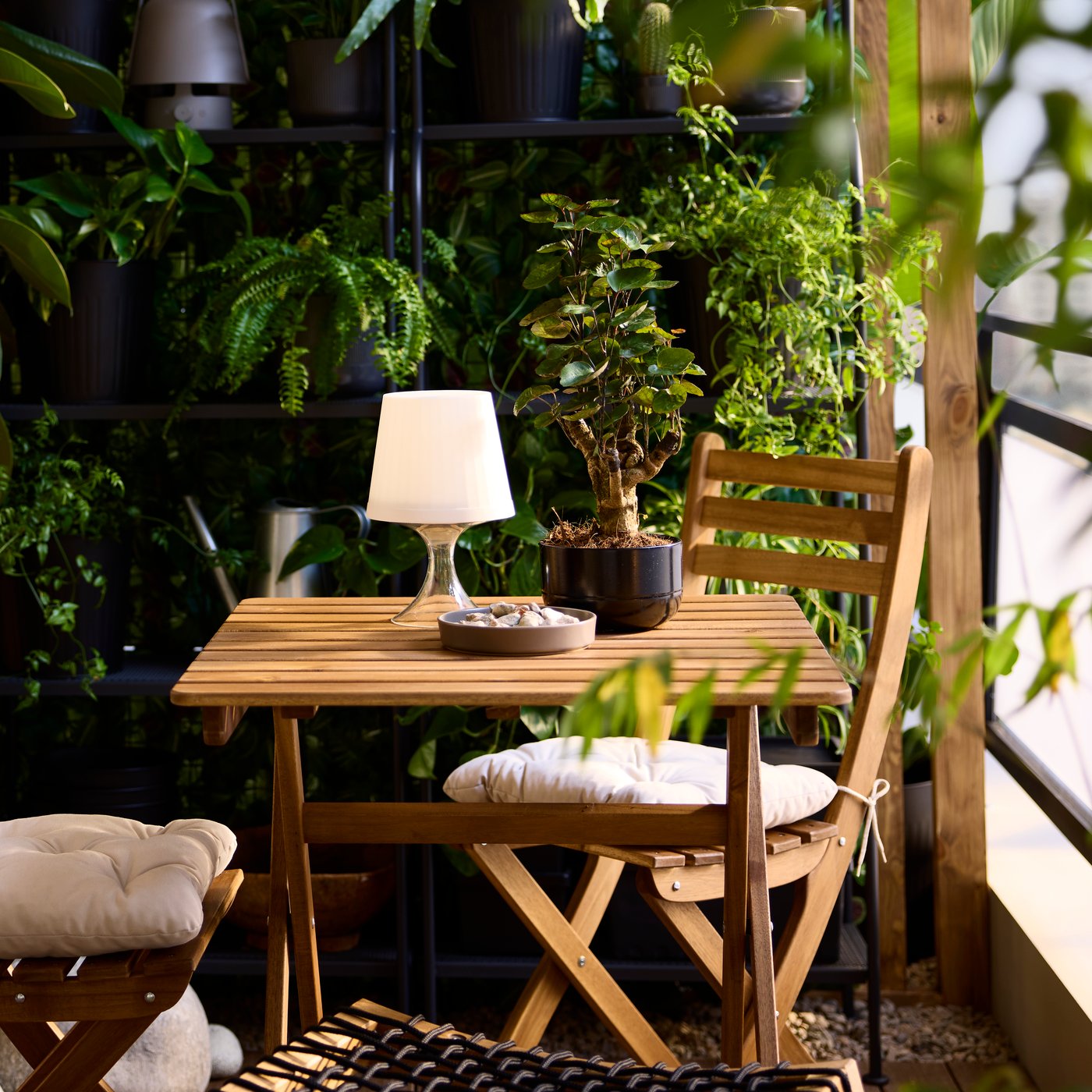 A light brown stained acacia ASKHOLMEN foldable table and 2 folding chairs are on a balcony with shelves and potted plants.