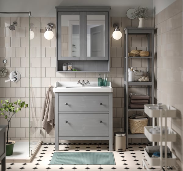 Un bagno HEMNES grigio visto frontalmente completo di lavabo in ceramica bianco, mobile per lavabo con due cassetti, pensile a specchio e mobile a colonna. Di fianco un box doccia e un carrello portatutto grigio.