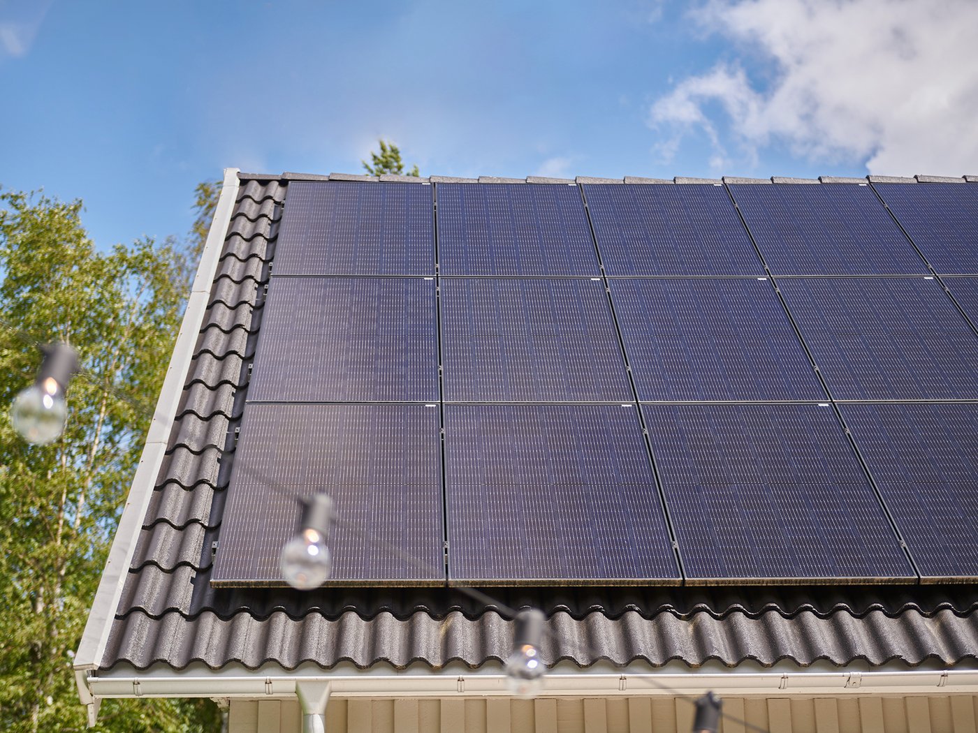 Solpaneler monterade på ett tak med mörkgrå takpannor, fotograferade i dagsljus mot en blå himmel med några moln. I förgrunden syns suddiga glödlampor som hänger på en ljusslinga, och i bakgrunden finns gröna träd.