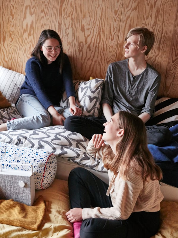 A teen room with a white SLÄKT bed frame, cushions and bed linen in different colors, and three friends hanging out.
