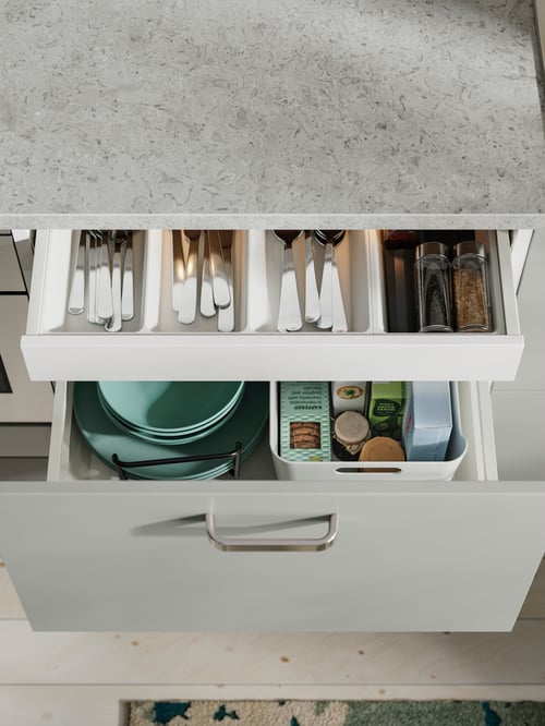 An opened drawer with a UPPDATERA spice rack placed inside and filled with spices is next to a set of cutlery, in a kitchen.