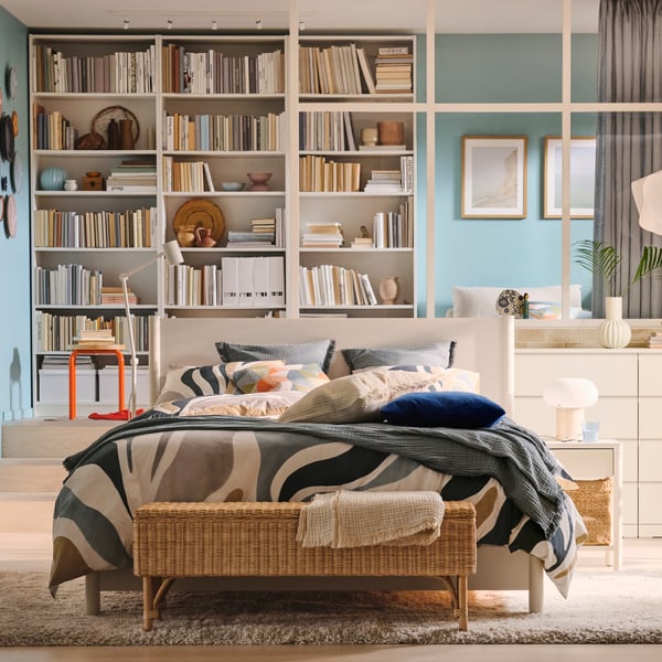 Una camera da letto con struttura letto con cassetti TONSTAD al centro con copripiumino e 2 federe  MYGGLASVINGE e una libreria BILLY in fondo alla stanza. La libreria è divisa da una struttura con assi bianche.
