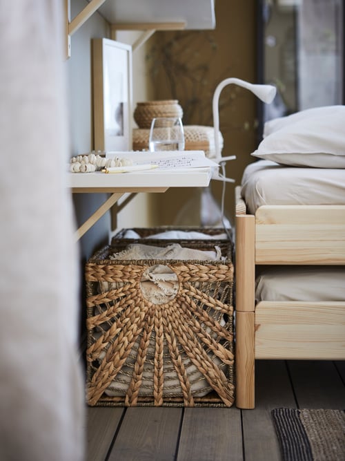 A shelf functioning as a bedside table, two LUSTIGKURRE natural water hyacinth/seagrass baskets and the head of a bed.