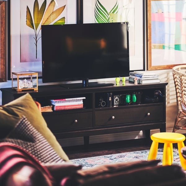 A black TV is on a dark brown stained IDANÄS TV bench, in front of which is a yellow MAMMUT children’s stool.