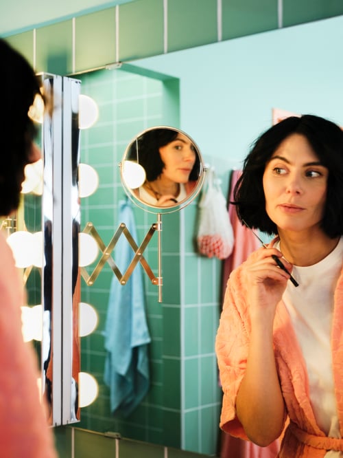 A woman holding a pair of scissors, looking into a positionable FRÄCK mirror while cutting her own hair in a green bathroom.