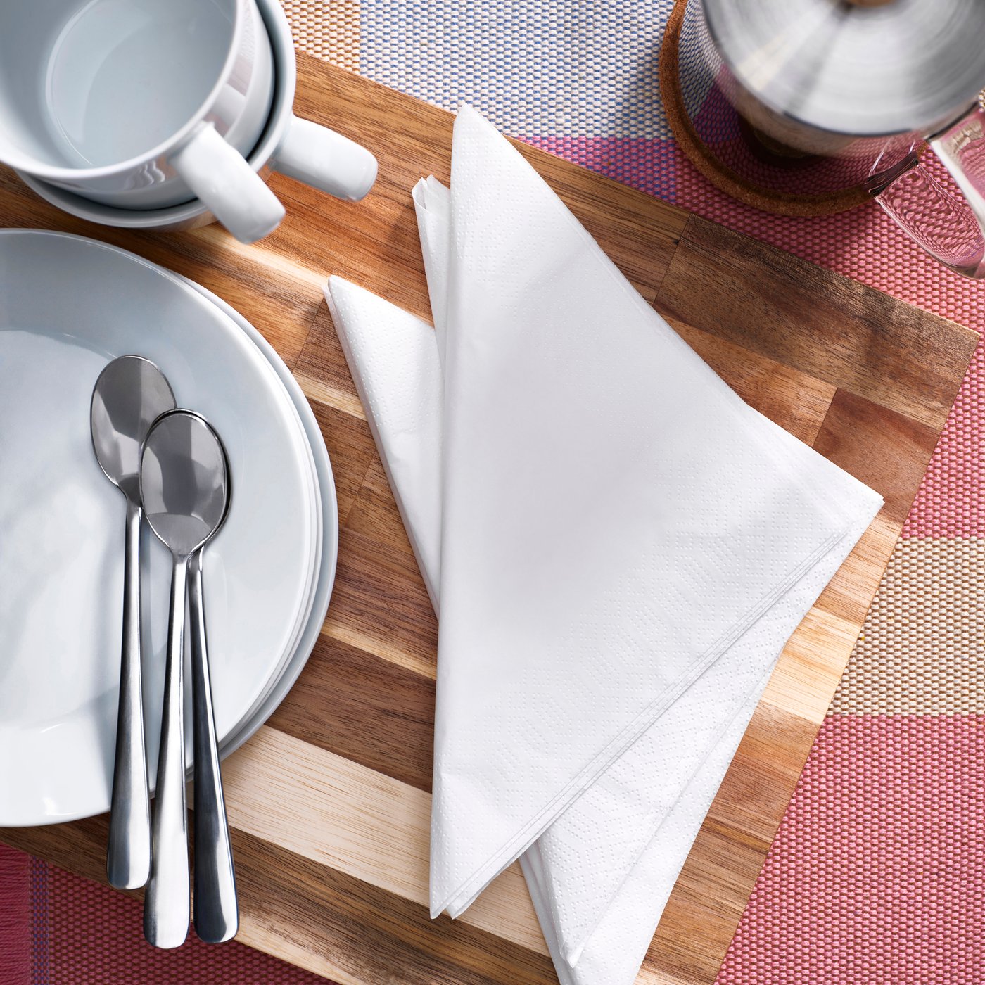 Dřevěné prkénko s bílými papírovými ubrousky, dvěma porcelánovými talíři, šálkem a třemi kovovými lžičkami. V pozadí barevný textilní ubrus a skleněná konvice s kovovým filtrem.