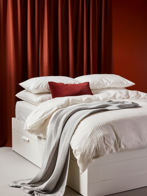 Bedroom with red walls, dark-red curtains, white bed frame with storage, white bed linens and grey throw.