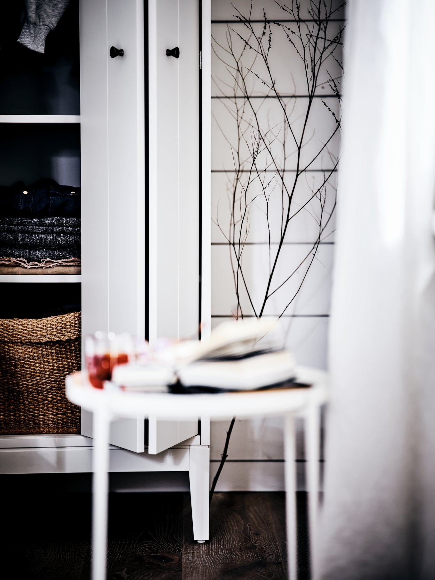 A white round table in front of a white open wardrobe with folding doors showing baskets and folded garments.