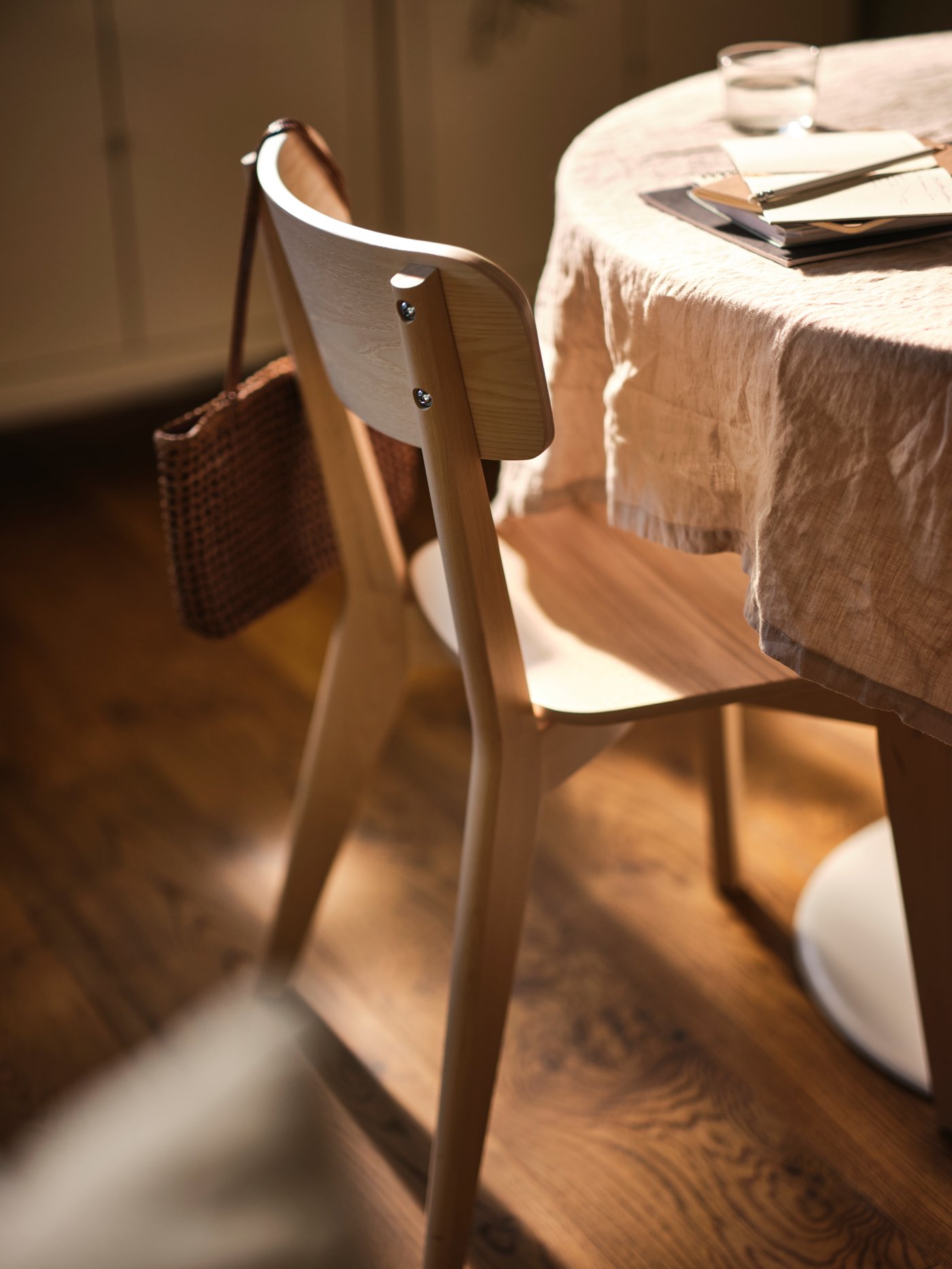 Una silla LISABO de fresno delante de una mesa cubierta con un mantel, con un bolso colgando del respaldo.