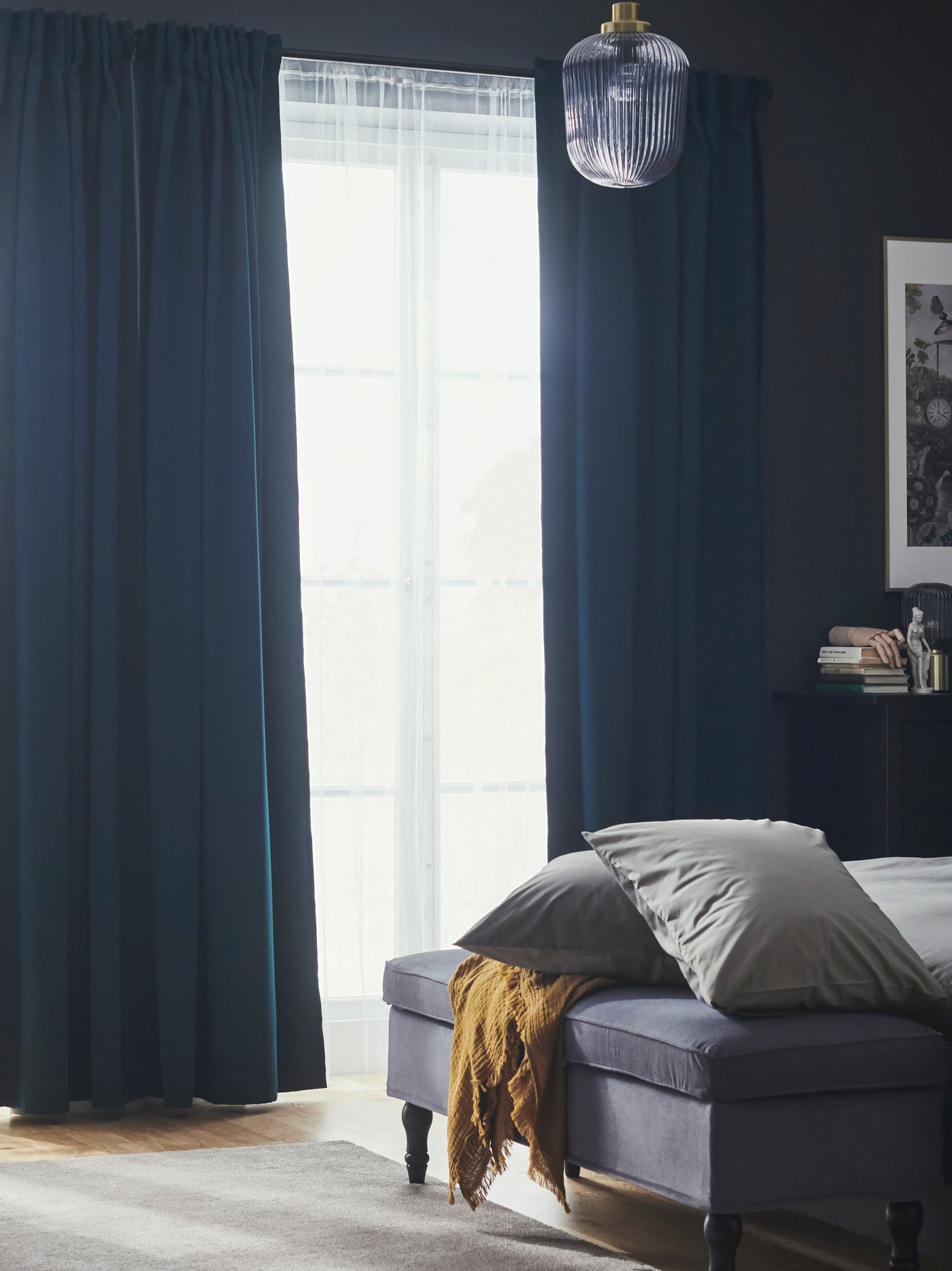 Un dormitorio con cortinas opacas MAJGULL de color turquesa oscuro y visillos en un gran ventanal frente a una cama y un banco.