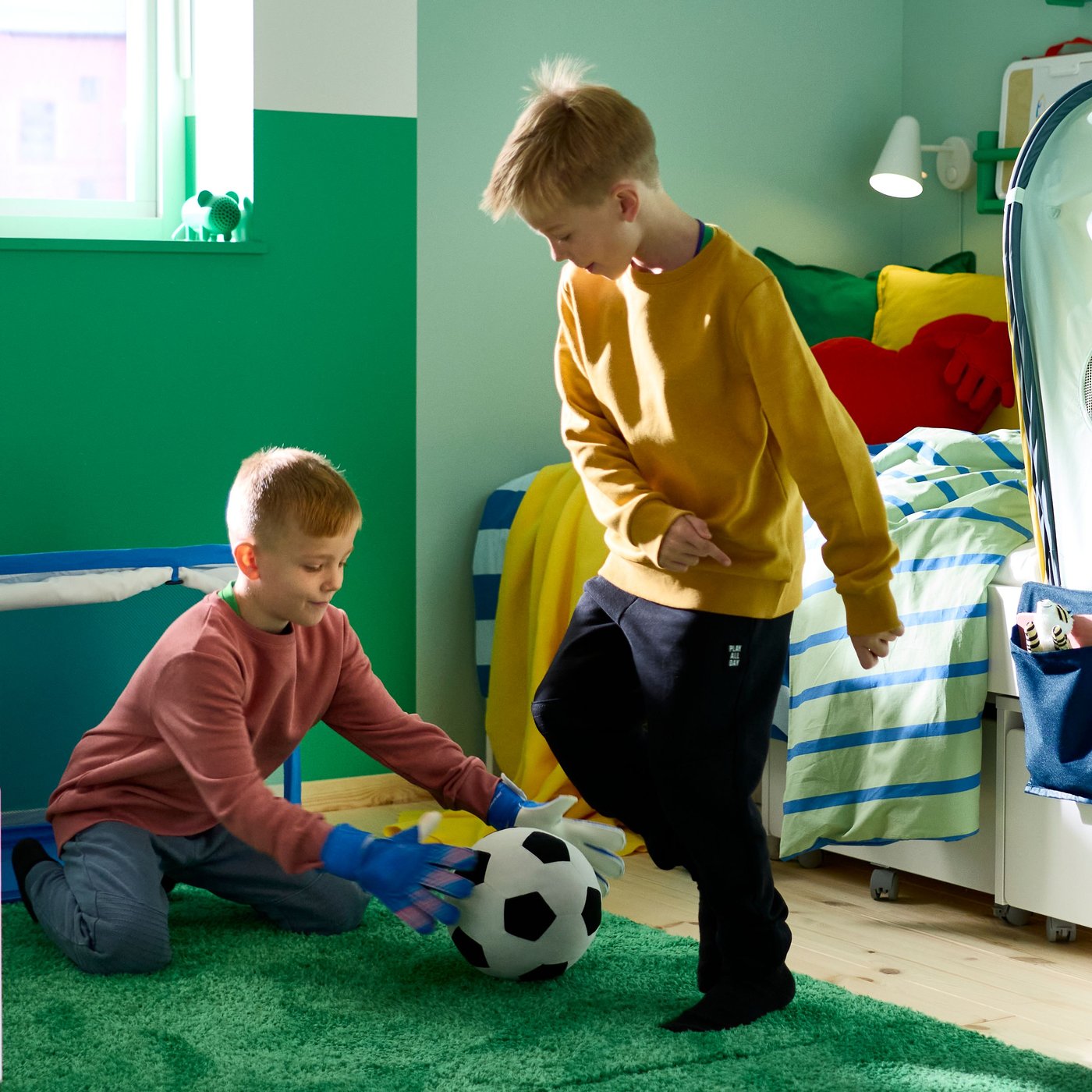 A green GREJSIMOJS portable Bluetooth speaker, shaped like a mouse, stands on a windowsill as two children play football.