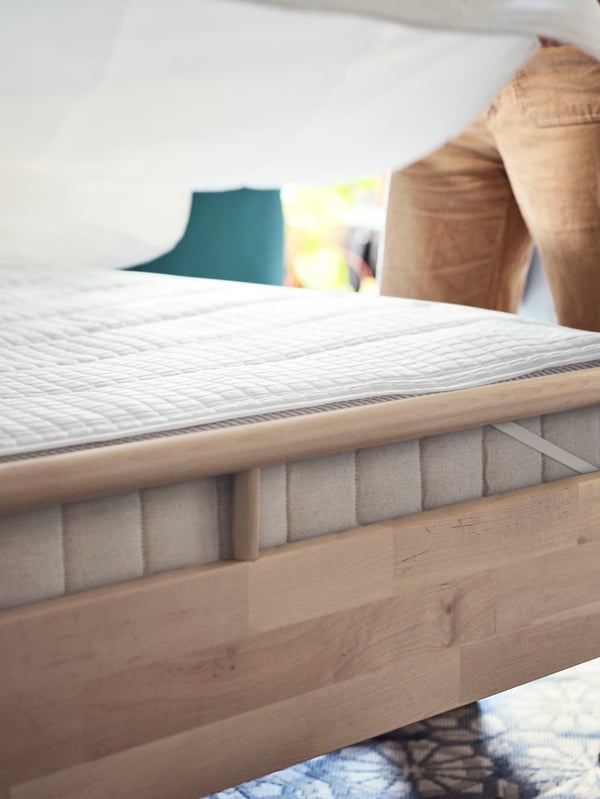 A birch bedframe with a thick beige mattress and white mattress protector. A man is making the bed with white bedding.