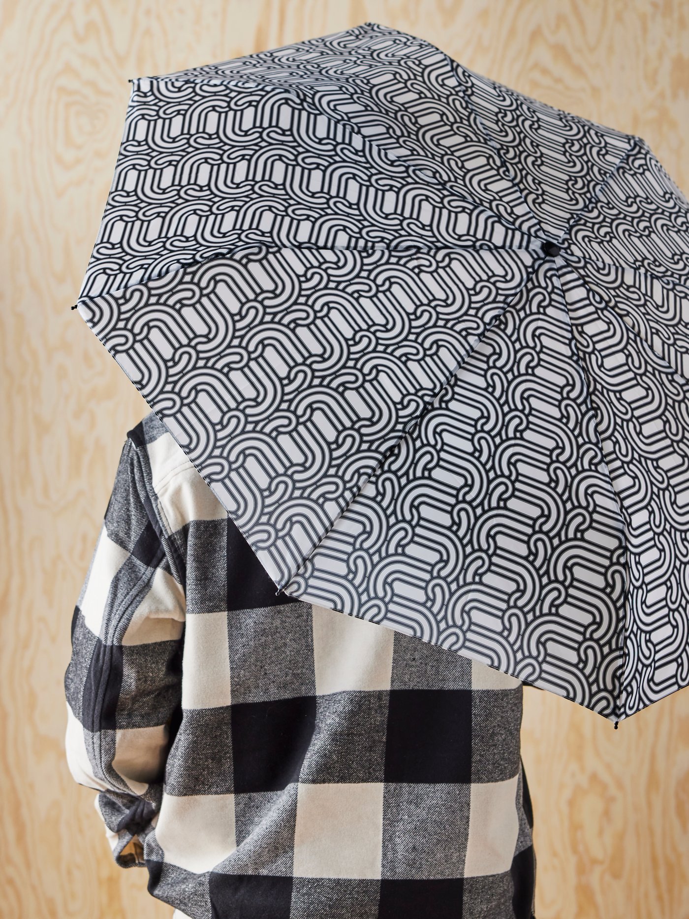 شخص يرتدي قميصاً بنقوش مربعات بالأبيض والأسود يمسك بمظلة SÖTRÖNN عليها نقوش مرسومة. 