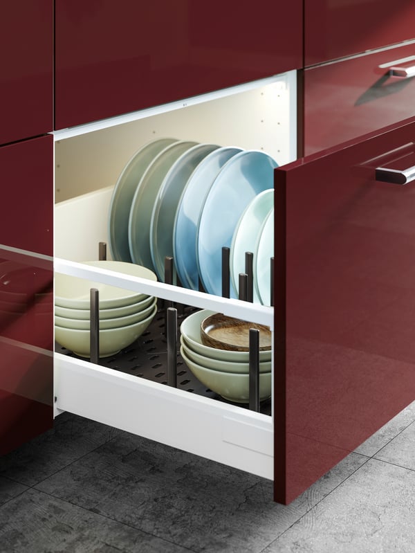 An open drawer in a kitchen with dark red-brown KALLARP fronts, showing plates and bowls on an UPPDATERA pegboard drawer organiser.