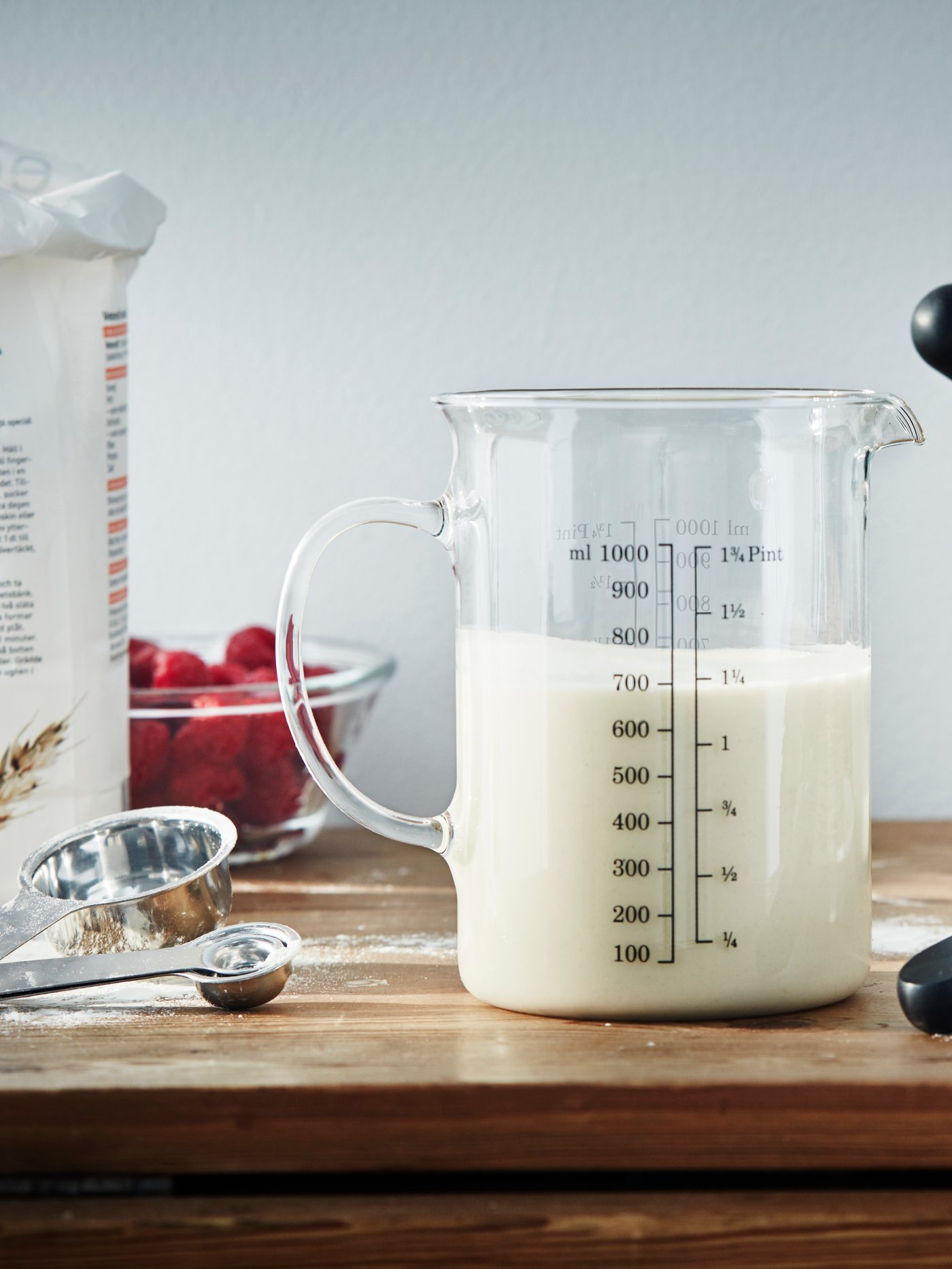 Odměrka VARDAGEN plná mléka na dřevěné desce zasypané trochou mouky. Nalevo jsou další odměrky VARDAGEN.