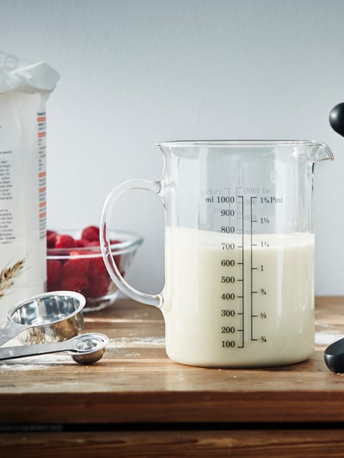 A measuring jug with milk beside a set of measuring cups, a bowl of berries and a bag of flour, on top of a wood countertop.