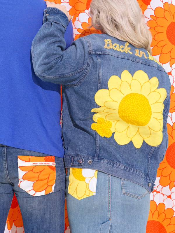 Twee personen gekleed in denim met een opvallende kontzak in het oranje en geel. De persoon aan de rechterkant heeft een SANDETERNELL spijkerjas aan met een gele bloem erop